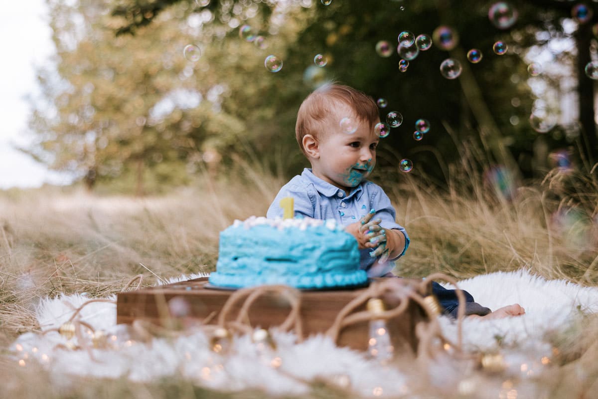 ein einjähriger junge sitzt beim cakesmash fotoshoot vor seiner torte und schaut sich seifenblasen an, die vor ihm im wind fliegen