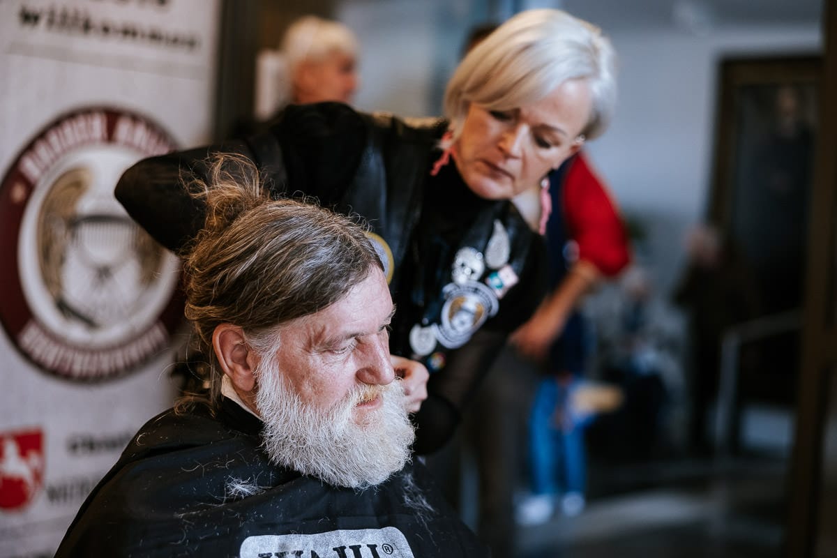 als fotografin bei den barber angels habe ich einen mann mit langen haaren fotografiert, die haare zum zopf zusammengebunden, dem eine friseurin den bart schneidet