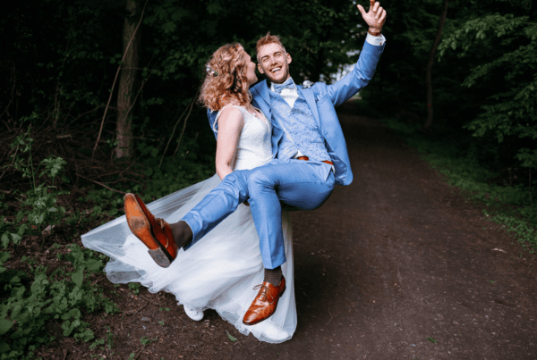 Braut trägt lachenden Bräurigam im Wald auf Händen.