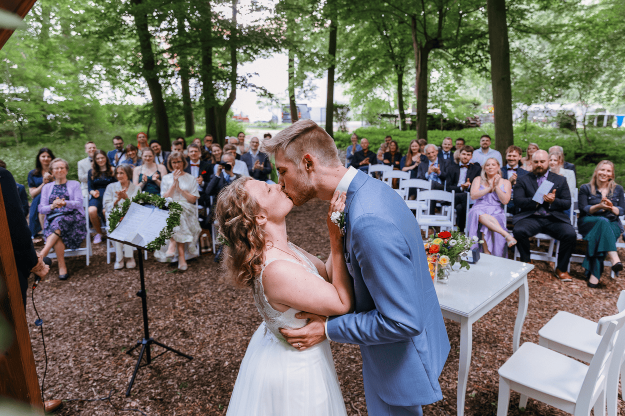 Hochzeitskuss im Wald mit Gästen hinter dem Waldhaus Ölper