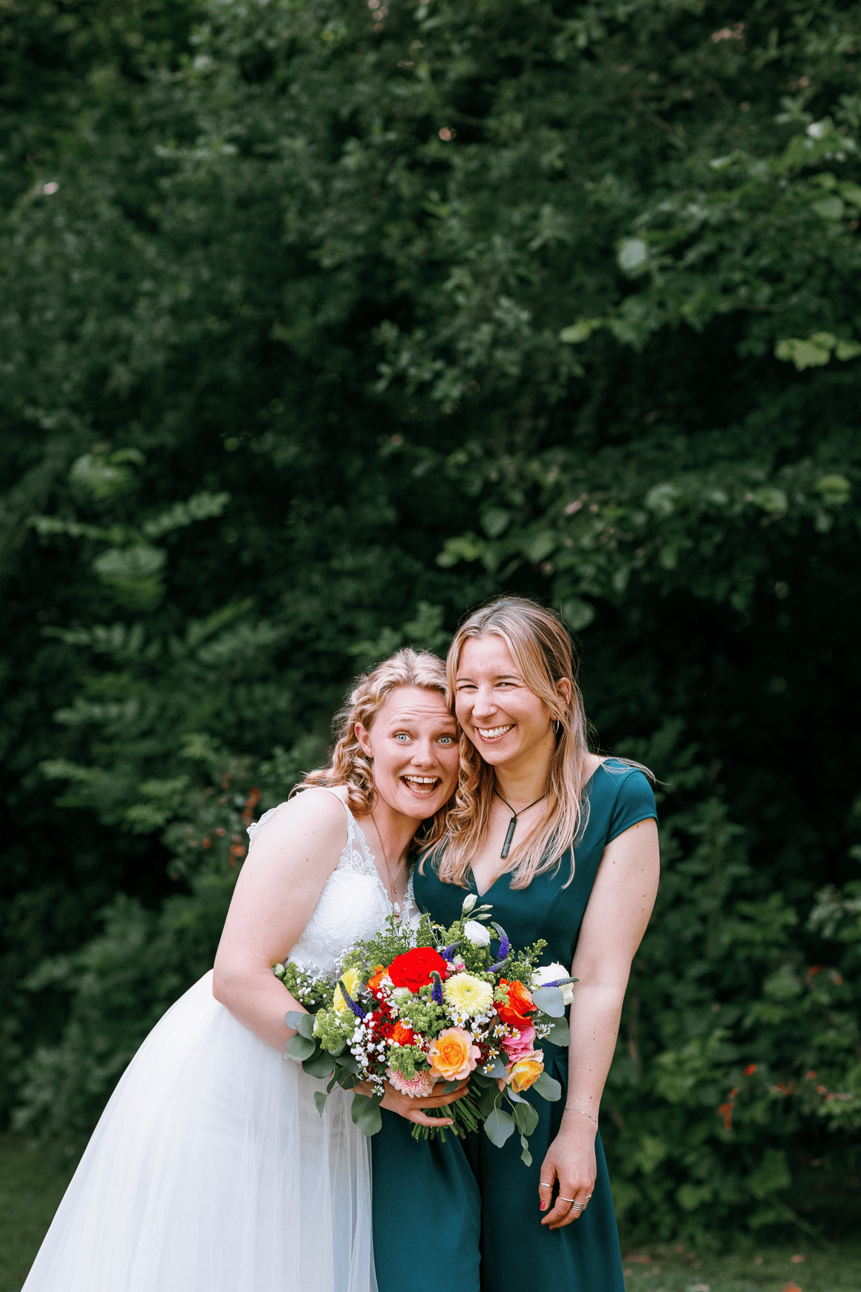 Braut und Freundin mit Brautstrauß vor Bäumen.