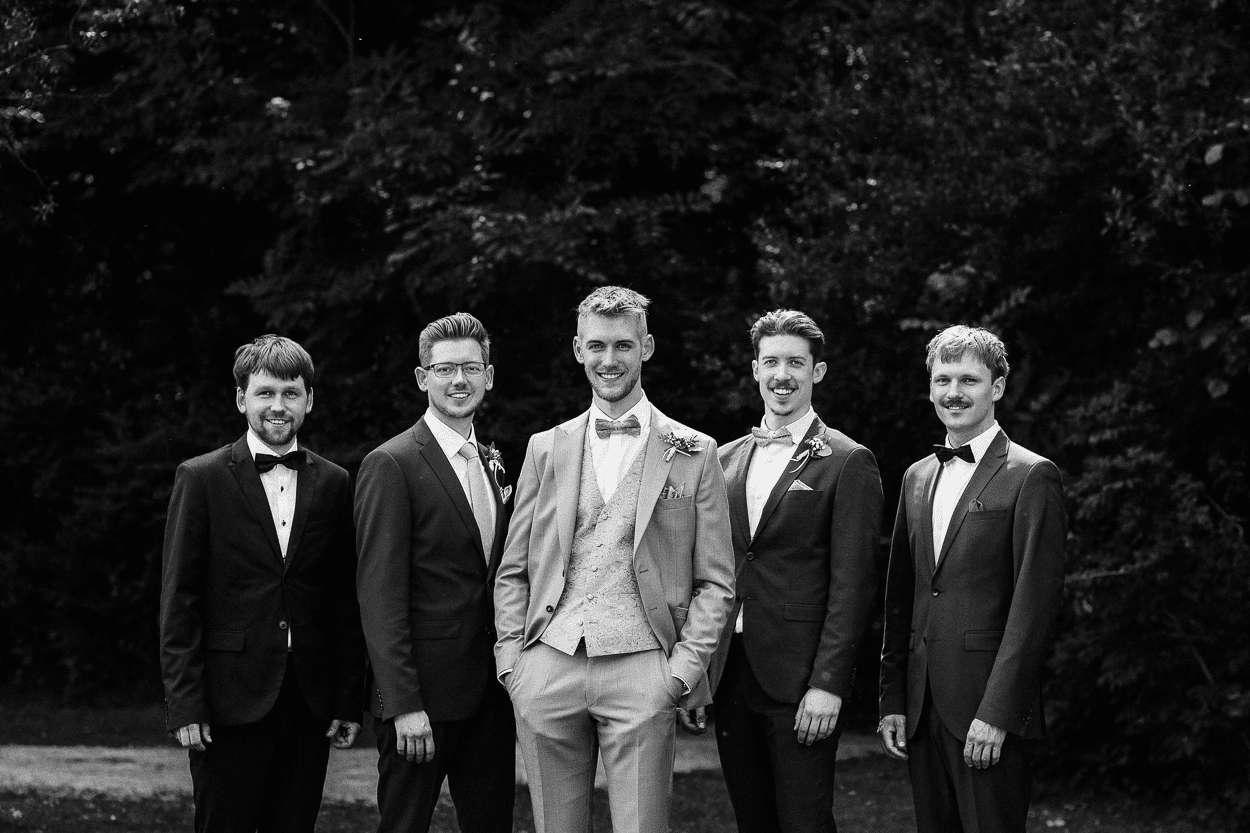 Bräutigam und vier Freunde in Anzügen, lachend im Freien.