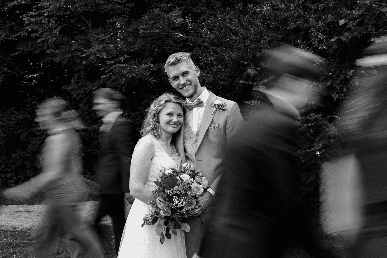 Brautpaar im Garten, verschwommene Gäste gehen am Paar vorbei.