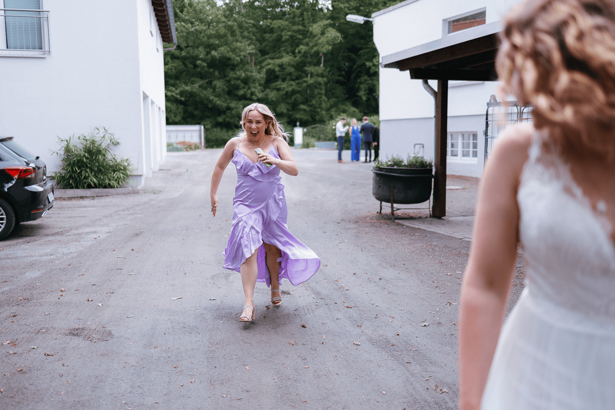 Lächelnde Frau in lila Kleid läuft auf eine Braut zu.