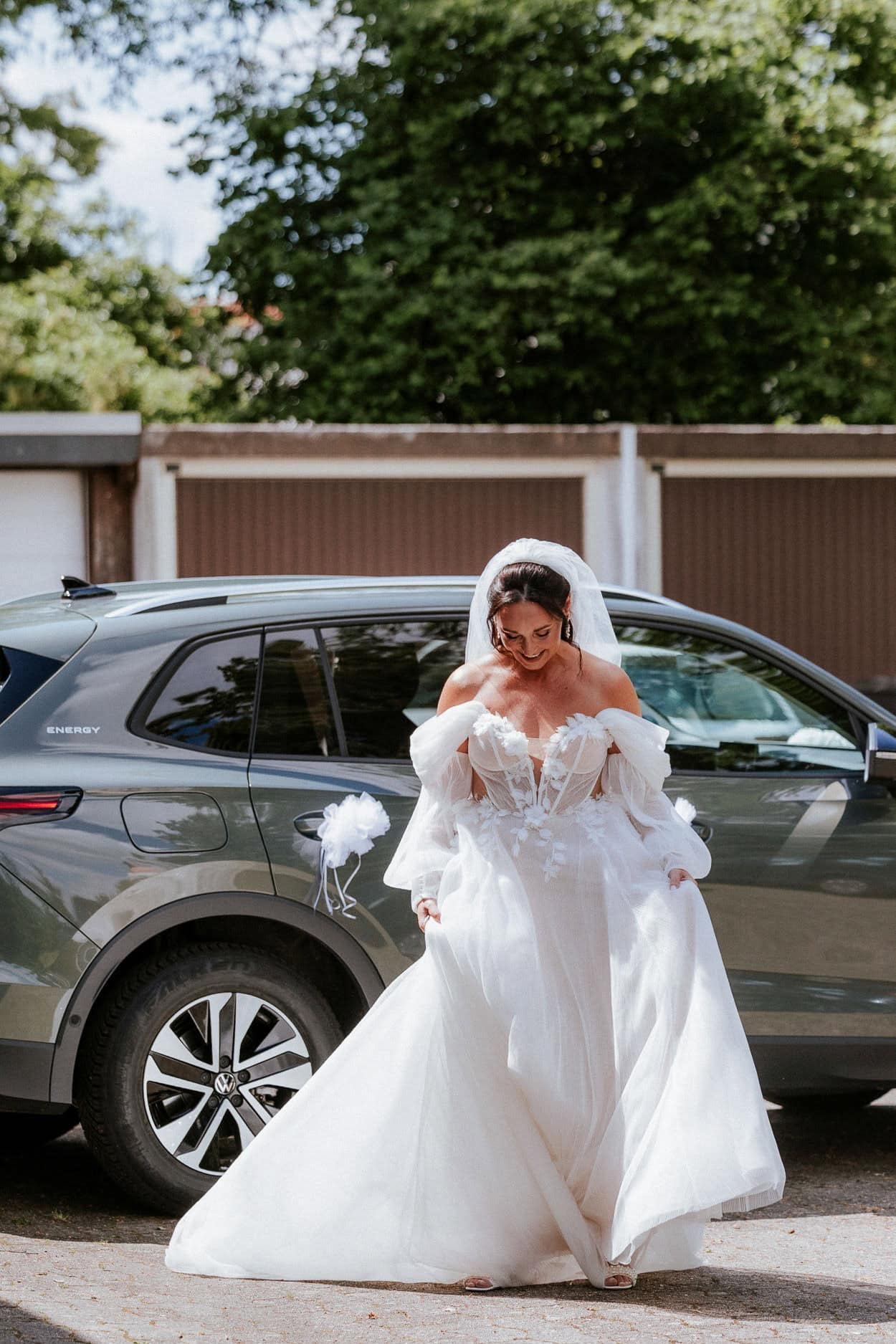 Braut im weißen Hochzeitskleid mit Puffärmeln steigt lächelnd aus dem Auto