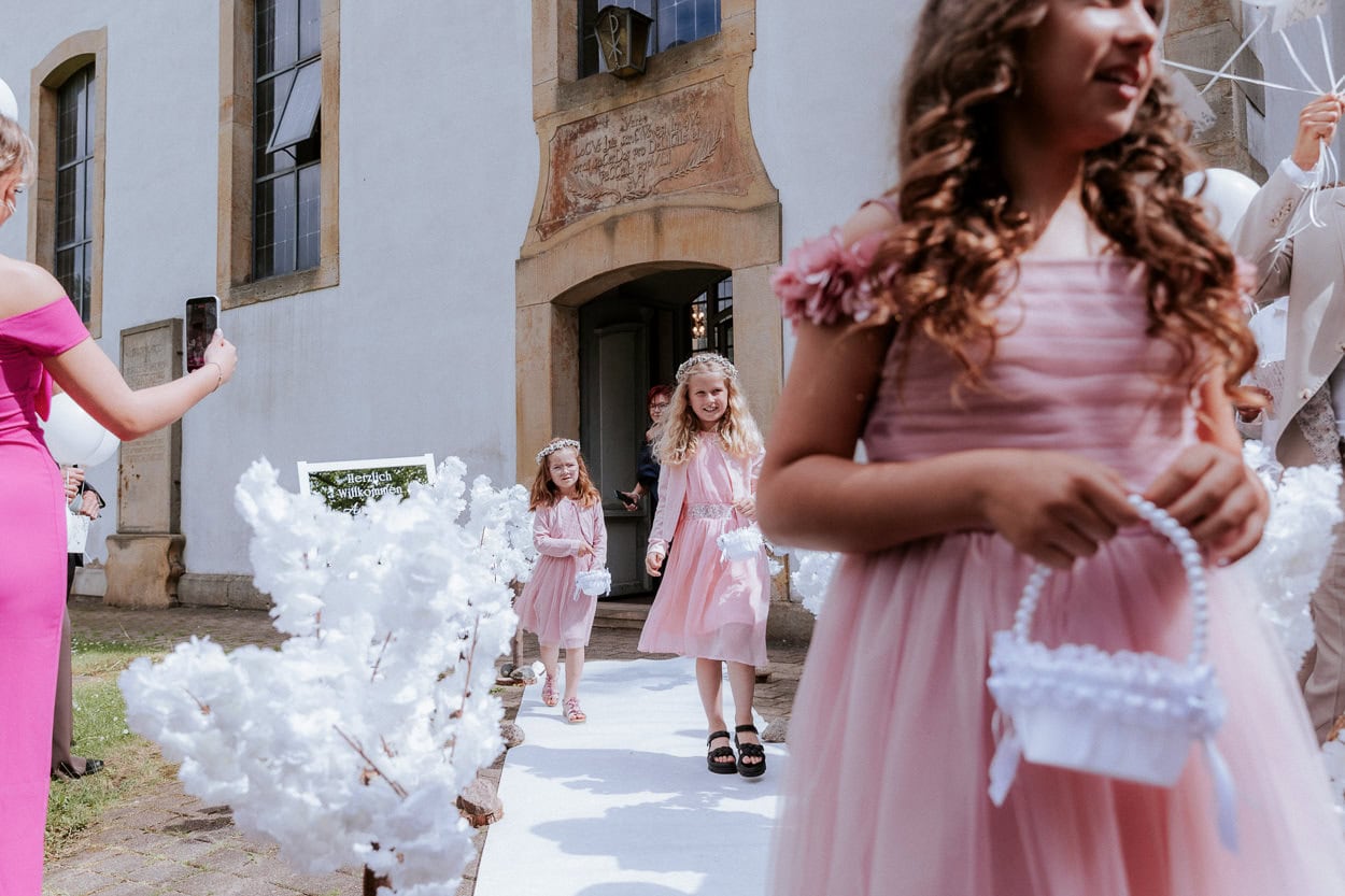 Blumenkinder in rosa Kleidern gehen den weißen Teppich vor der Kirche entlang