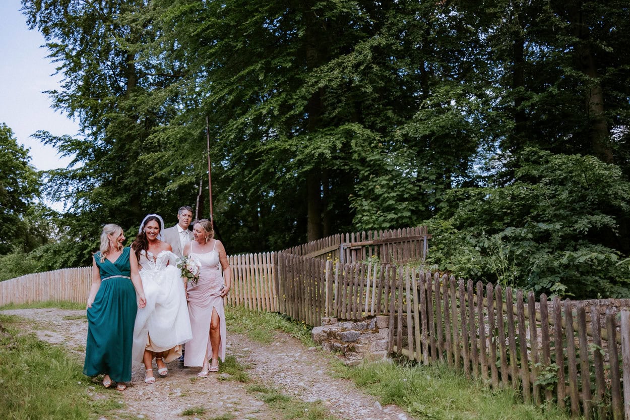 Braut geht lachende mit zwei Freundinnen und dem Bräutigam einen Weg entlang, gesäumt von Holzzäunen