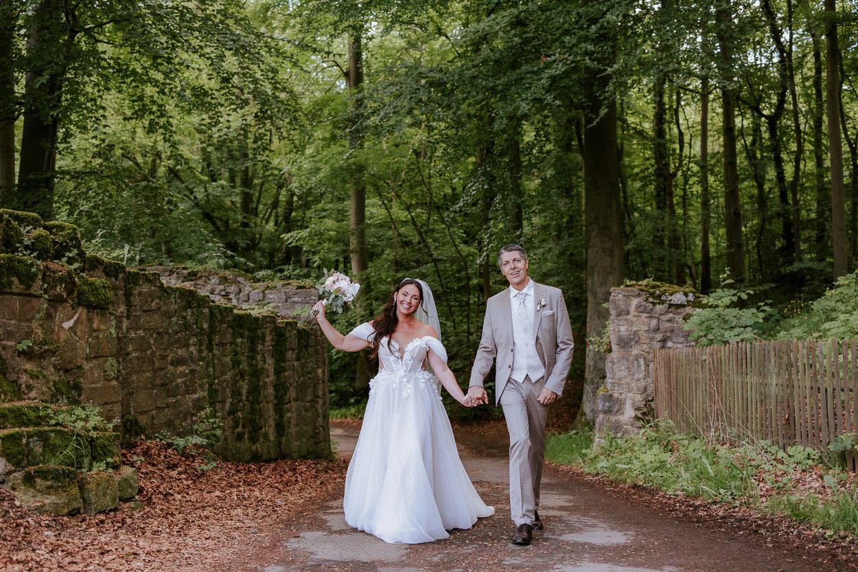 Brautpaar geht händchenhaltend lachend durch eine alte Mauerruine im Wald auf die Fotografin zu