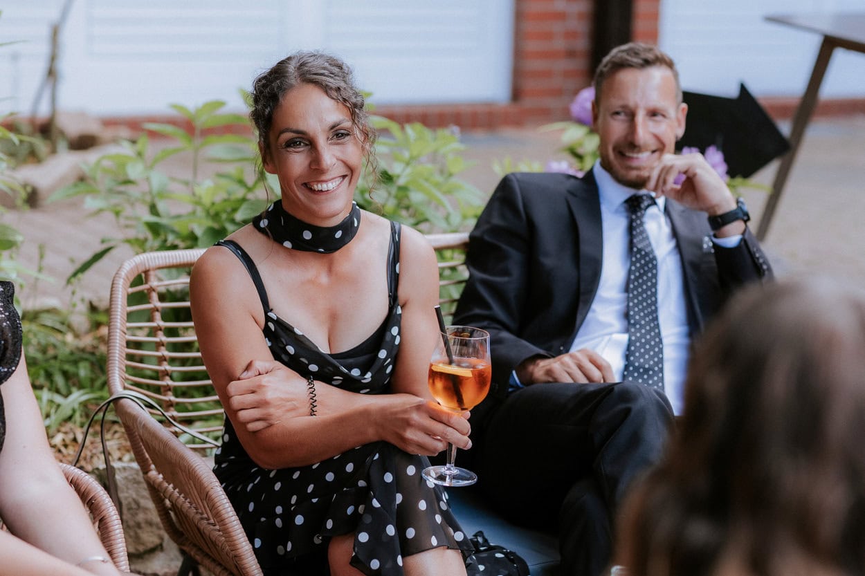 Lächelnde Hochzeitsgästin mit Cocktail sitzt neben einem Gast im Anzug