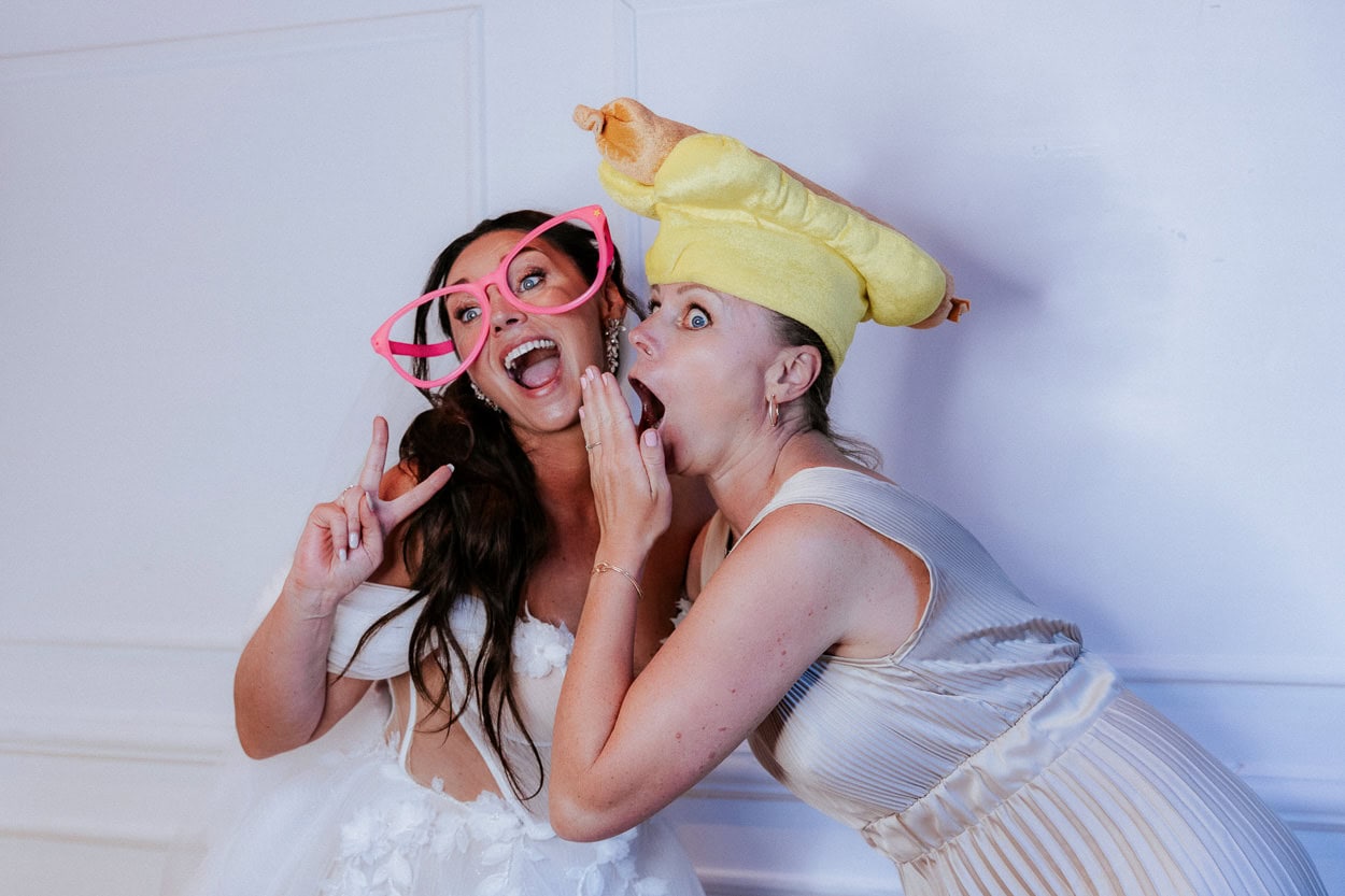 Braut mit riesiger pinker Fotobox-Brille und Gästin mit Hotdoghut lachen ausgelassen
