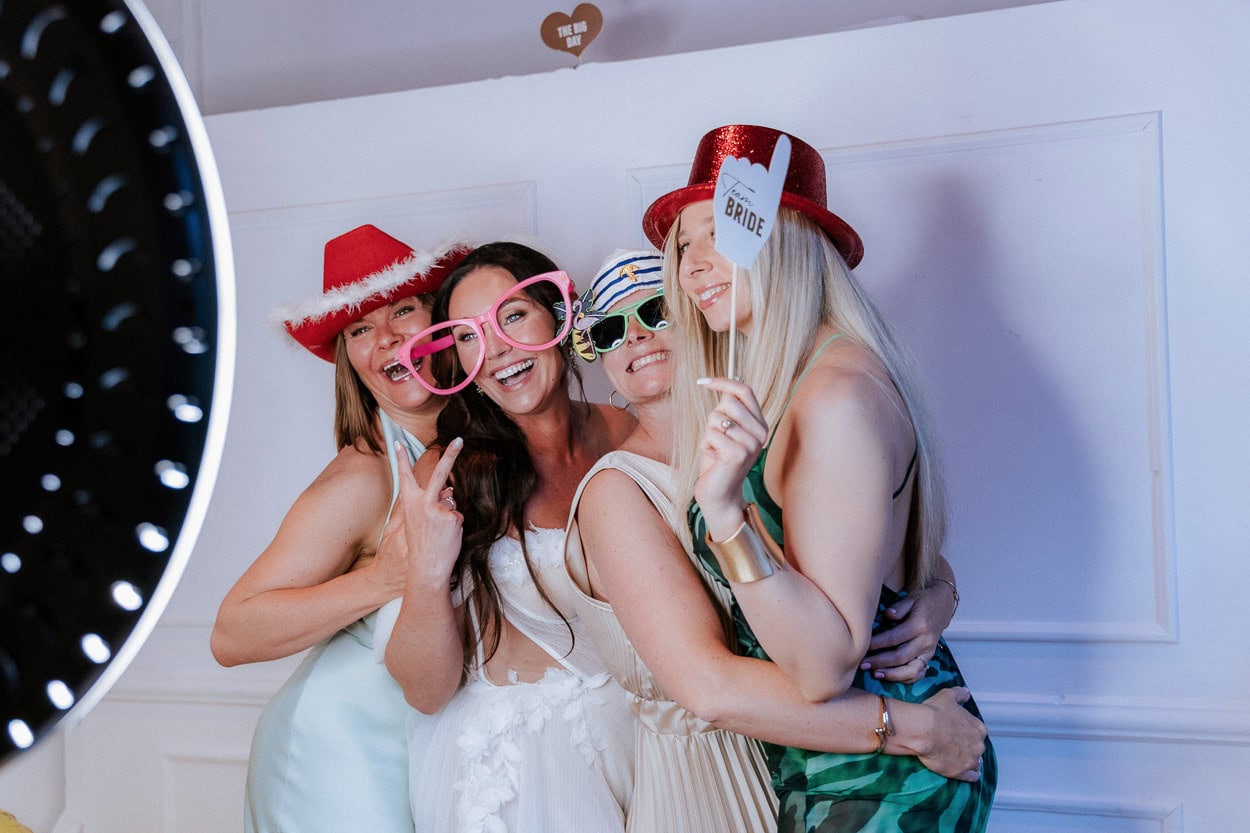 Braut posiert lachend mit drei Freundinnen in bunten Fotobox-Accessoires