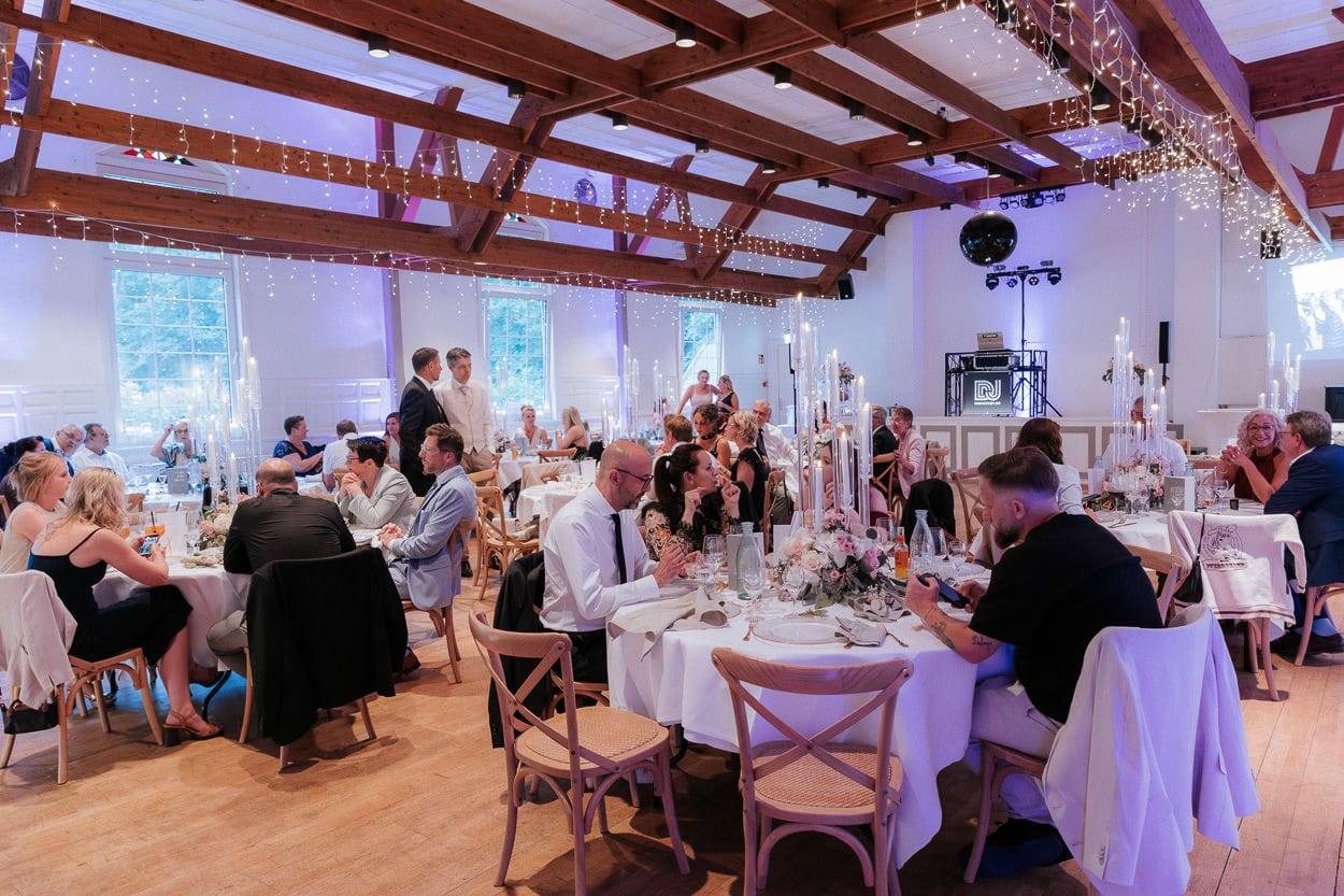 Festlich beleuchteter Saal mit Hochzeitsgesellschaft an Tischen, DJ-Pult im Hintergrund